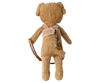 A Maileg Puppy Supply, Harness stands upright, facing away. It wears a cozy harness with a small pouch attached and a brown object that appears to be a bow slung over one shoulder. The bear's fur is light brown and shaggy.