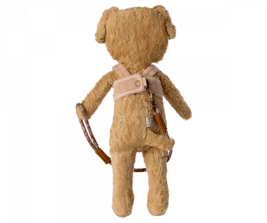 A Maileg Puppy Supply, Harness stands upright, facing away. It wears a cozy harness with a small pouch attached and a brown object that appears to be a bow slung over one shoulder. The bear's fur is light brown and shaggy.