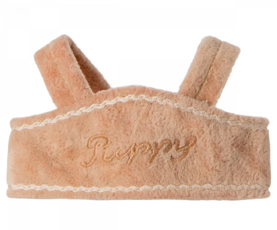 A soft, pinkish-beige Maileg Puppy Supply, Harness with "Puppy" embroidered on it in cursive beige thread. The velvety harness features decorative white stitching along the edges and two padded shoulder straps.