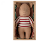 A Maileg Pig In Box, Baby - Girl, wearing a red and white striped shirt, lies inside a rectangular wooden box. The box is lined with a patterned fabric resembling a small blanket and pillow. The playset features the pig's simple, smiling face, with its head slightly turned to the right.