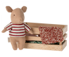 A small, Maileg Pig In Box, Baby - Girl wearing a red and white striped sweater stands next to a wooden box. Inside the box, there are pieces of patterned fabric, one of which is draped over the edge, creating an adorable playset scene that could easily charm both adults and children alike.