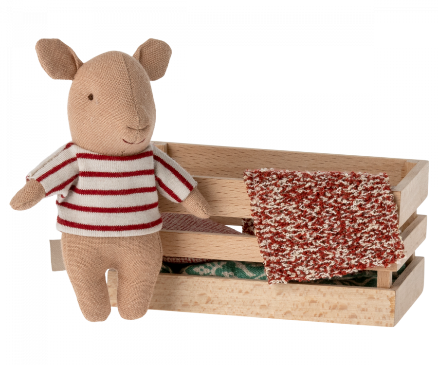 A small, Maileg Pig In Box, Baby - Girl wearing a red and white striped sweater stands next to a wooden box. Inside the box, there are pieces of patterned fabric, one of which is draped over the edge, creating an adorable playset scene that could easily charm both adults and children alike.