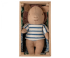 A Maileg Pig In Box, Baby - Boy with a striped blue and white shirt lies in a wooden box, resting on a patterned cushion. The bear's expression is neutral.