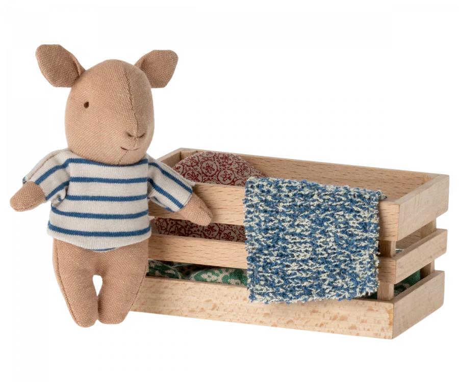 A Maileg Pig In Box, Baby - Boy toy standing next to a small wooden box filled with assorted colorful fabrics, isolated against a white background.