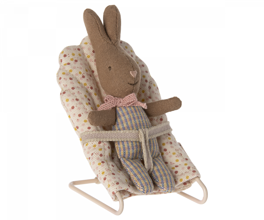 A Maileg My Rabbit - Rose/Blue, possibly the smallest bunny you've seen, wearing striped overalls and a pink bow tie sits in a baby bouncer. The bouncer features a light-colored, polka-dotted cushion and metal frame, with the bunny secured by a fabric strap around its waist—truly one of the must-have accessories for any nursery table.