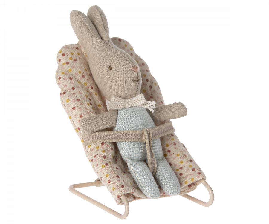 The Maileg My Rabbit - Blue, praised as the smallest bunny, is dressed in a checkered blue outfit and a white bowtie. It sits comfortably in a padded, patterned baby bouncer featuring a floral design on a white background. The rabbit is secured by a simple cloth harness—an adorable addition to any nursery table.