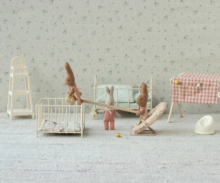 A cozy nursery scene features three Maileg My Rabbit - Dots fabric toys in a soft pastel setting. One is nestled in a crib, another playfully balances on a seesaw, and the third relaxes in a bouncer. The arrangement is completed with a high chair, changing table, and toy potty adorned with delightful accessories.