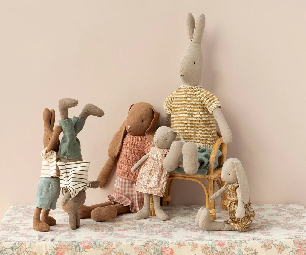 A group of Maileg Bunny, Size 2 - Flower Dress dolls made from soft natural fabrics is gathered together. Two small bunnies are doing handstands on the left, while two medium-sized bunnies in dresses stand in the center. A large bunny in striped clothes, crafted from recycled polyester, sits on a chair to the right. They all have a cozy, handmade look.