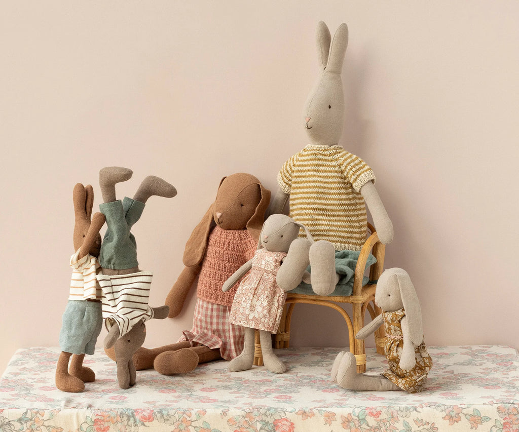 A group of Maileg Bunnies, Size 1 - Classic Flower Dress, some dressed in striped outfits and others in floral dresses made from soft natural fabrics, are arranged on a floral-covered table. One Maileg Bunny, Size 1 - Classic Flower Dress is seated in a wicker chair, two are standing on their hands, and another is sitting on the edge of the table.