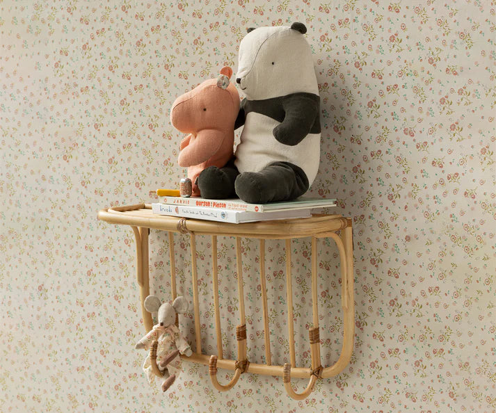 A shelf with wooden hooks is mounted on a floral wallpapered wall. On the shelf are two stuffed animals, a pink rhinoceros and a Maileg Panda Stuffed Animal, Medium from the Maileg Safari Friends collection, sitting on top of several children's books. A small mouse plush is hanging from one of the hooks below.