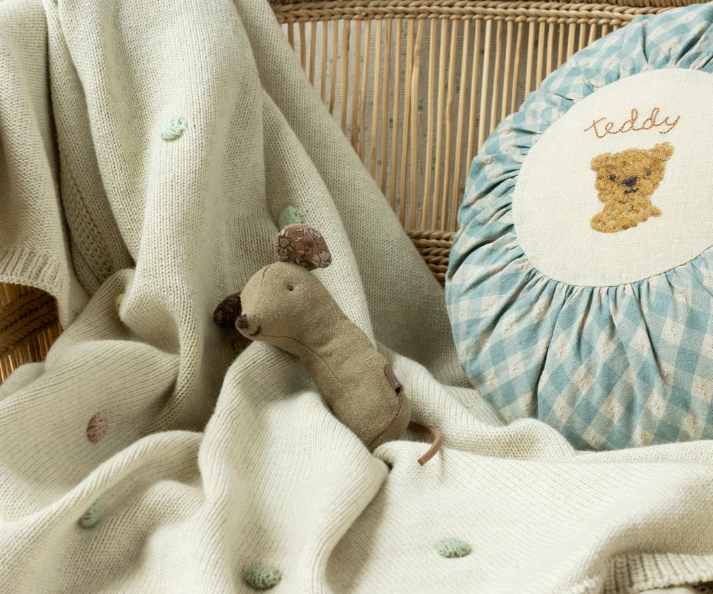 A cozy setup with a soft, knitted blanket draped over a wicker chair, adorned with small pastel-colored dots. A Maileg Lullaby Friend Rattles, Mouse Rattle in Light Umber lies on the blanket, and a round cushion with a teddy bear design and the word "teddy" sits beside it.