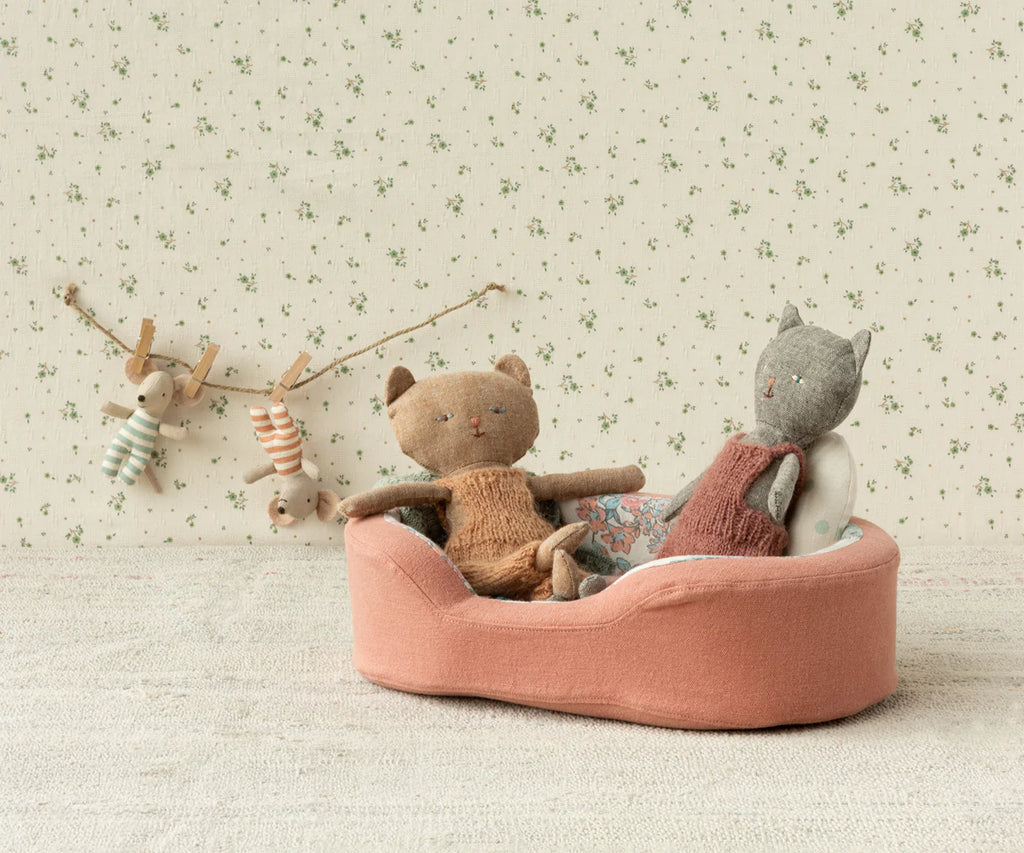 Two fabric cats, dressed in knitted clothes, sit in the Maileg Small Cozy Bed - Coral, cushioned with a soft blanket. Behind them, a string hangs with two small stuffed rabbits in striped outfits, attached with clothespins. The background wallpaper is beige with a subtle floral pattern.