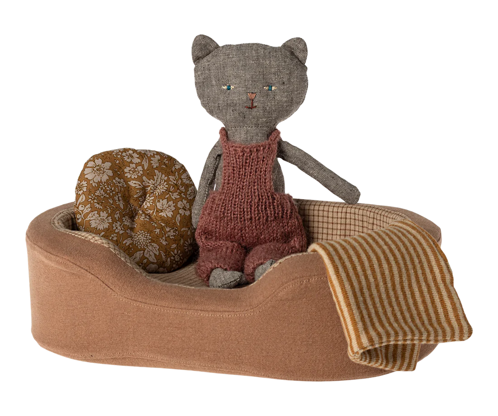 A handmade fabric cat doll wearing a red knitted overall sits in a Maileg Cozy Bed. The small pet basket includes a round, floral-patterned cushion and a soft blanket with yellow and white stripes draped over the side.