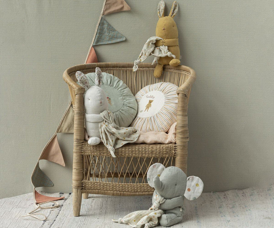 A wicker chair with plush toys sits against a light gray wall. The chair holds the Maileg Lullaby Friends - Bunny - Dusty Yellow (Plays Music) and a round pillow featuring a giraffe illustration. An elephant plush toy sits on the floor, ready to join its lullaby friends. Triangular fabric bunting hangs in the background for decoration.