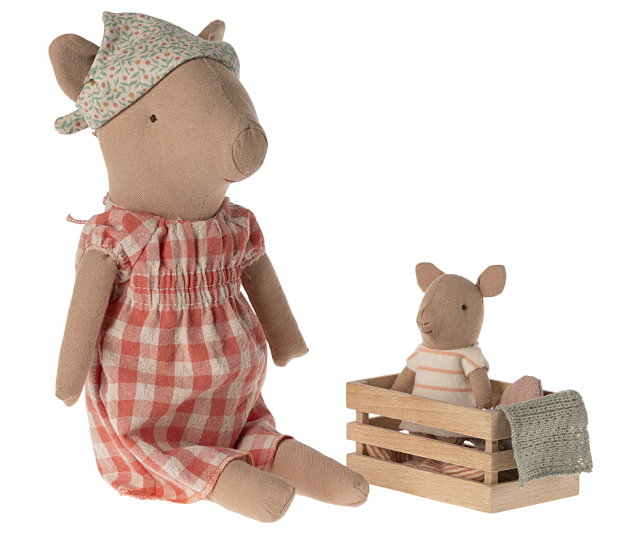 Two Maileg Pig - Girl toys are shown. The larger girl pig wears a floral headscarf and a red-checkered dress, sitting with legs extended. The smaller pig, dressed in a striped shirt, sits in a small wooden crate that is partially covered with fabric. They make up the sweetest pig family scene.