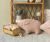 A cozy room with a small yellow doll bed, where a small stuffed pink pig made of soft fabric lies under a striped blanket. Nearby is another larger pink stuffed pig, specifically the Maileg Polly Pork, Medium. On a shelf, a green stuffed animal sits above some books. Decor includes flowers, strawberries, and macarons. Some toys feature recycled polyester filling for eco-friendliness.