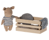 A small tan plush toy resembling a bear is wearing a white and blue striped shirt and standing beside a wooden box. The box contains a Maileg Pig In Box, Baby Boy - Blue, a circular blue-patterned cushion, and soft bedlinen.