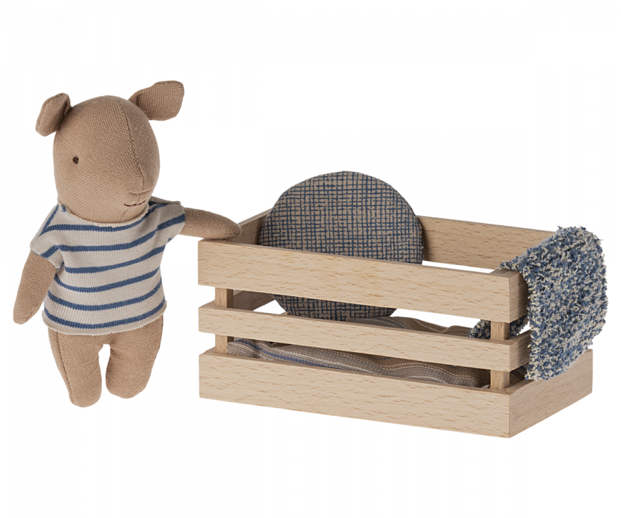 A small tan plush toy resembling a bear is wearing a white and blue striped shirt and standing beside a wooden box. The box contains a Maileg Pig In Box, Baby Boy - Blue, a circular blue-patterned cushion, and soft bedlinen.