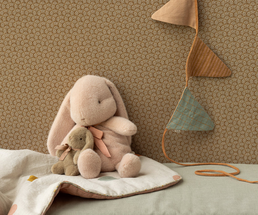 A cozy scene features two Maileg Small Plush Bunny - Powder toys made with polyester fillings on a soft blanket, set against a patterned wallpaper. A string of triangular flags in earthy tones hangs decoratively in the corner, adding charm to the setting.