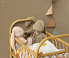 Two Maileg Medium Plush Bunnies in Dusty Brown, crafted from soft fabric with polyester fillings, sit in a wicker crib against a patterned brown wall. The crib is lined with a white sheet featuring colorful leaf designs, and a string of triangle flags hangs nearby, enhancing the charming scene.