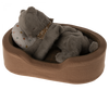 A Maileg Plush Kitten, Earth grey with a brown floral-patterned ribbon around its neck is curled up, resting its front paws on a polka-dotted pillow in a cosy basket. The plush bed has a checked pattern on the interior.