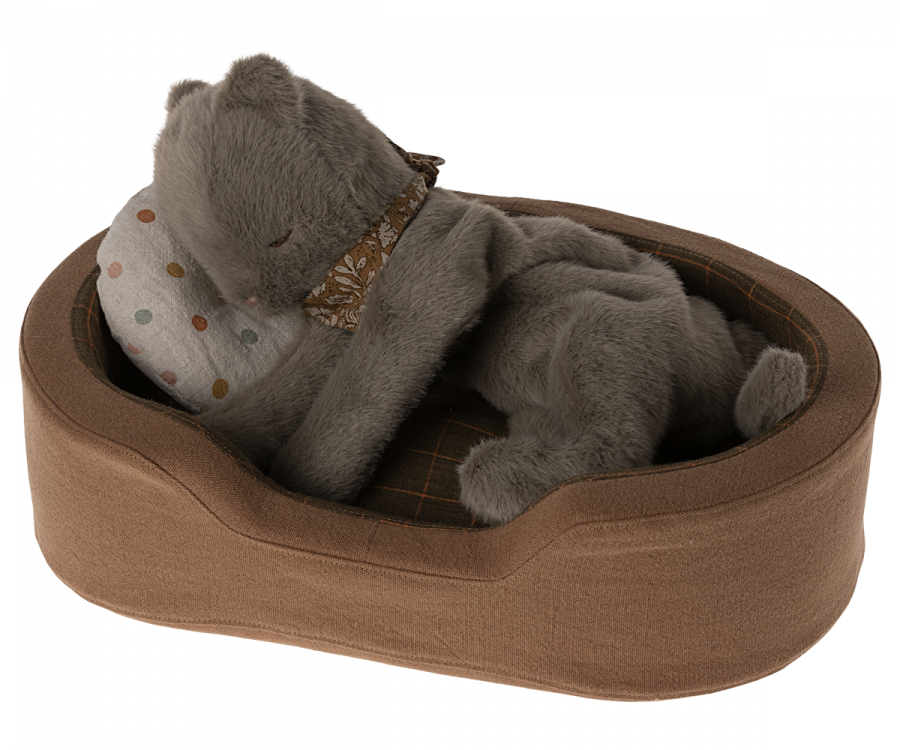 A Maileg Plush Kitten, Earth grey with a brown floral-patterned ribbon around its neck is curled up, resting its front paws on a polka-dotted pillow in a cosy basket. The plush bed has a checked pattern on the interior.