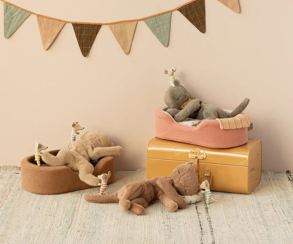 Four Maileg Plush Kittens, Cream peach (ships in approximately one week), two grey and two brown, each holding a small toy giraffe, are posed on various objects including a pink cushion, a brown basket, and a yellow box. In the background, a string of colorful triangular flags is strung across the wall.