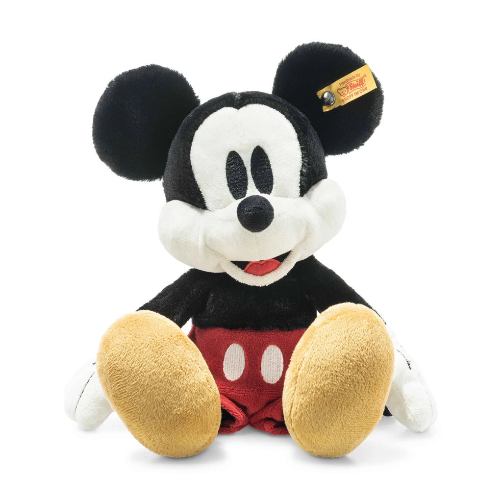 This Steiff, Disney's Mickey Mouse Stuffed Plush Toy, 12 Inches, with Steiff craftsmanship, features the iconic black ears and head, a white face, red shorts adorned with two white buttons, and large yellow shoes. The cheerful toy is smiling and has a tag attached to one ear.