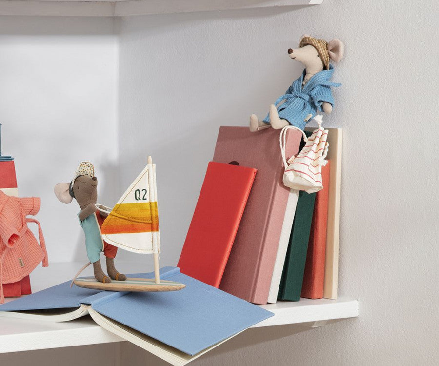 Two small stuffed mice are displayed on white shelves with books. One mouse, dressed in a blue robe and sunglasses, sits on top of stacked books, while the other, wearing a mint dress, stands on a toy sailboat. ***Maileg Extra Clothing: Bathrobe - Dusty Blue*** is also draped over the books.
