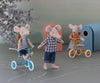 Three toy mice are depicted in a whimsical setting, two of which are riding bicycles. One mouse in a blue checkered shirt from the Maileg Extra Clothing: Clothes For Dad Mouse set stands in the forefront while the other two, dressed in striped shirts, ride bicycles. A soft, pastel backdrop and small birdhouse are in the background.