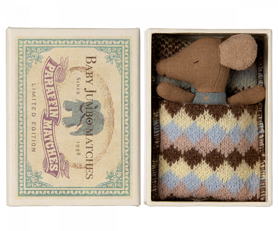 A small baby mouse with a brown body and large ears lies in a cozy, patterned sleeping bag inside a matchbox labeled "Maileg Sleepy Wakey Baby Mouse In Matchbox - Blue" by Maileg. The box is open, revealing the mouse snugly tucked in.