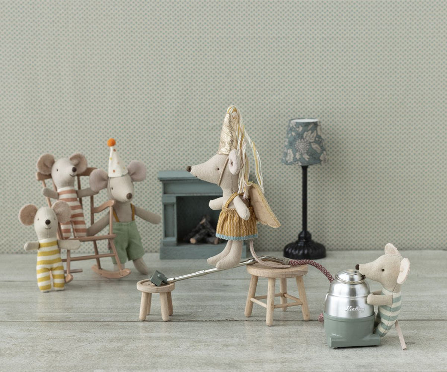 A whimsical scene featuring small toy mice in a cozy room. One mouse wearing *Maileg Fairy clothes In Suitcase, Little Sister Mouse* and a party hat is on a seesaw, while another controls it with a vacuum cleaner. In the background, three other mice are on a rocking horse. Nearby, *Maileg Fairy clothes In Suitcase, Little Sister Mouse* peeks from behind a tiny fireplace and lamp.