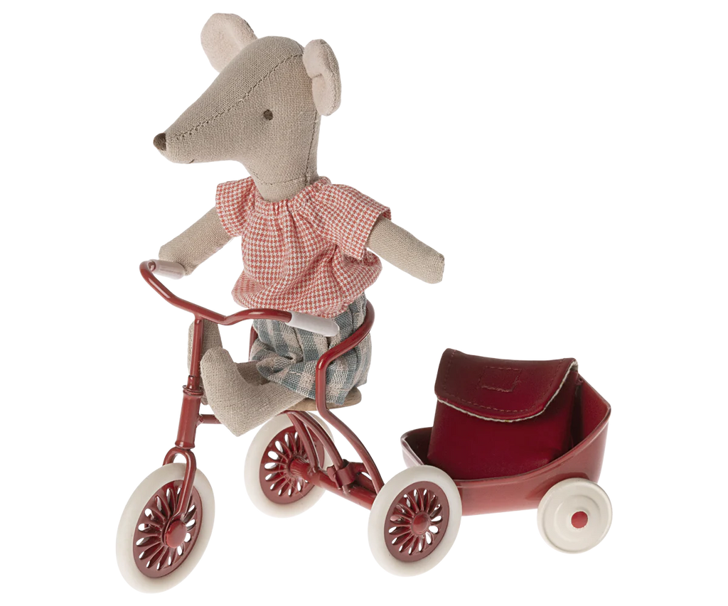 A Maileg Big Sister With Backpack - Red wearing a red-checked shirt and blue shorts sits on a red tricycle, gripping the tricycle handles with confidence. A small red trailer is attached at the back. The toy's fabric has a textured, handmade appearance, perfect for kindergarten playtime adventures.