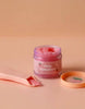 An open jar of Tiny Tummies - Strawberry jelly food from Tiny Harlow is on display, accompanied by a pink magic spoon. The jar, designed in pink and purple hues, has its lid placed nearby on a light brown surface.