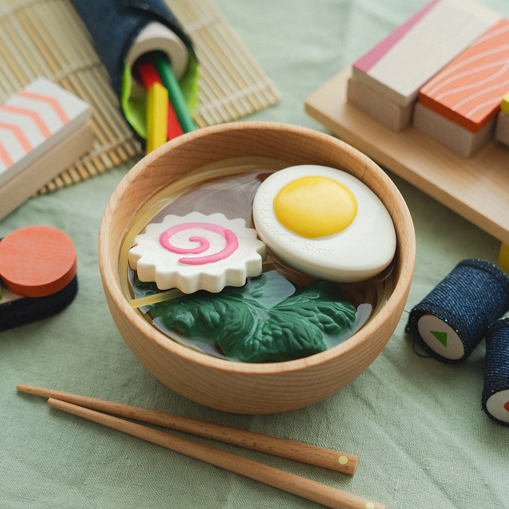 A Ramen Bowl Playset (ships in approximately one week) with toy food items resembling ramen ingredients, including an egg, narutomaki, and vegetables. Surrounding the bowl are wooden toy sushi pieces, non-toxic toy noodles in a fabric case, and a rolled bamboo mat on a green fabric surface—perfect for sensory play noodles.