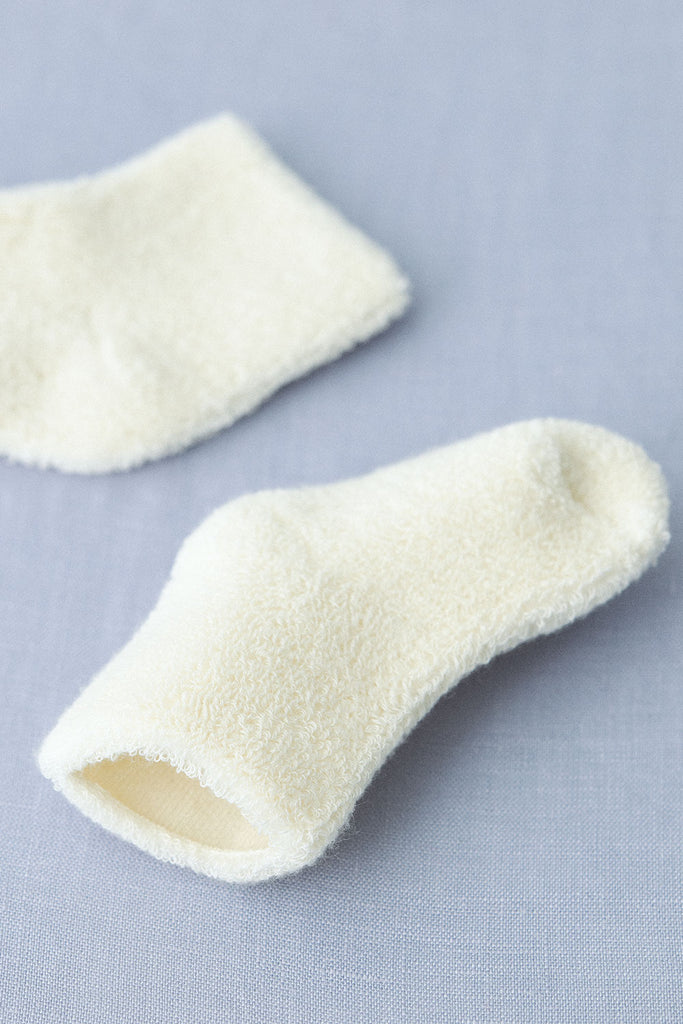 A pair of small, white, fluffy Baby Pile Knit Socks - Natural are placed on a light blue fabric surface. One sock is positioned flat, revealing its opening, while the other is slightly rolled, emphasizing its soft and textured material. These adorable Lithuania baby accessories add a touch of cozy charm to any outfit.