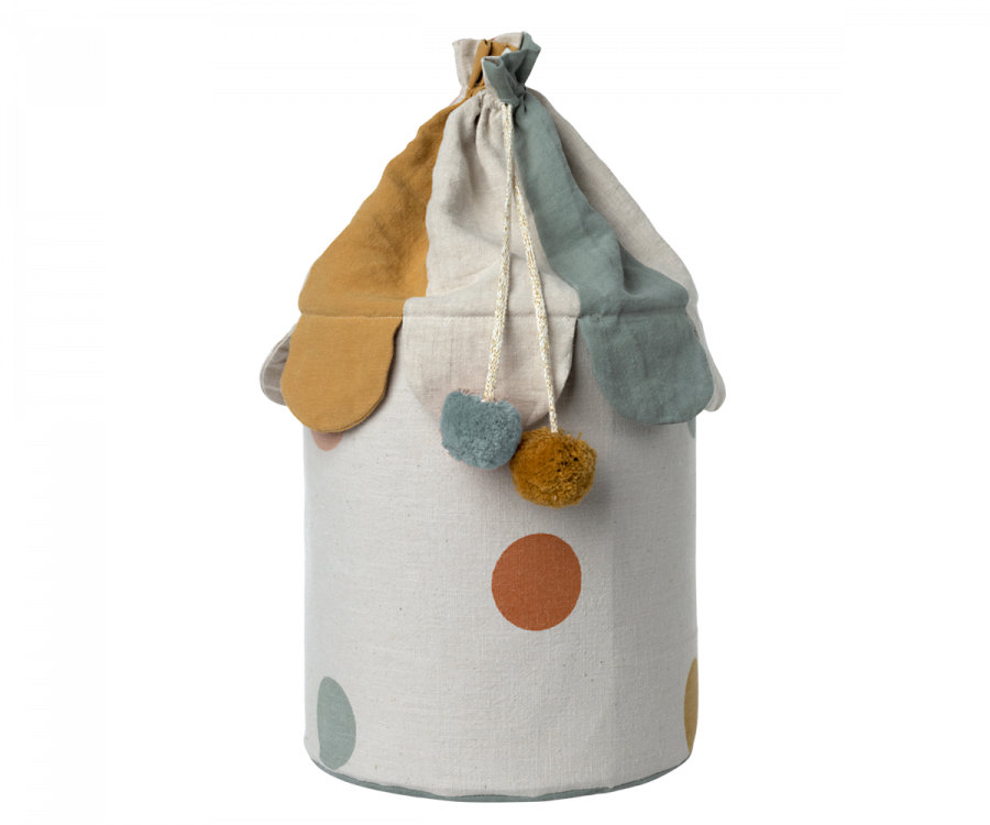 The Maileg Basket, made from cotton linen and designed in the shape of a circus tent, showcases a multicolored scalloped top adorned with pom-pom accents and features a convenient drawstring closure. Ideal for toy storage, it has a white base decorated with large colored circles.