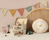 A cozy scene featuring the Maileg 2 Piece Suitcase Set, with one open showcasing small stuffed animals in hats. Nearby are a miniature rocking chair, a green toy car, a tiny rug, a small lamp, and a round cushion. A colorful banner of Maileg fabrics hangs above, while a wicker backdrop completes the setting.