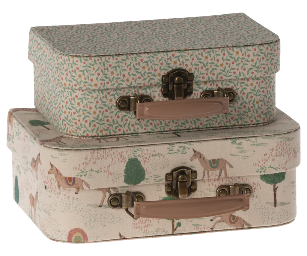 Two decorative suitcases from the Maileg 2 Piece Suitcase Set are stacked. The top suitcase, crafted from Maileg fabrics, features a floral pattern with green leaves and red dots, while the bottom one displays a whimsical design with horses, trees, and rainbows. Both suitcases have beige handles and antique-style metal clasps—perfect for stylish storage.