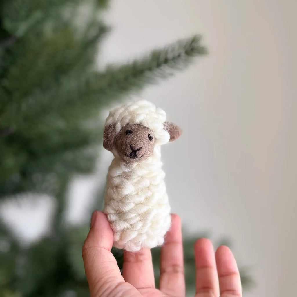 A hand holds the Fluffy Sheep Finger Puppet, a small knitted creation handmade in Nepal, in front of a blurred green foliage background. The cream-colored sheep, crafted from soft, chunky yarn, features detailed brown facial accents.