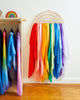 In the playroom, a Sarah's Silk Large Playsilk Display - Rainbow made from Red Oak MDF gracefully holds vibrant fabric streamers in red, orange, yellow, green, blue, and purple against a white wall. To the left, a wooden coat rack showcases more colorful garments while a playful rainbow decoration sits on top.