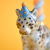 A collectible keepsake, the Jaguar Cake Topper is an exquisite fine porcelain figurine featuring a cheetah adorned with a small blue hat decorated with orange polka dots. Set against a solid bright yellow backdrop, the cheetah showcases detailed and delicate decorative markings.