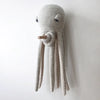 A BigStuffed Octopus - Big Original, evocative of an underwater world, is mounted on a wall. The fabric of this stuffed octopus is textured and off-white, and its tentacles hang down loosely. With large droopy eyes, the soft toy appears to have a wooden rod protruding from its mouth. This item ships in approximately one week.