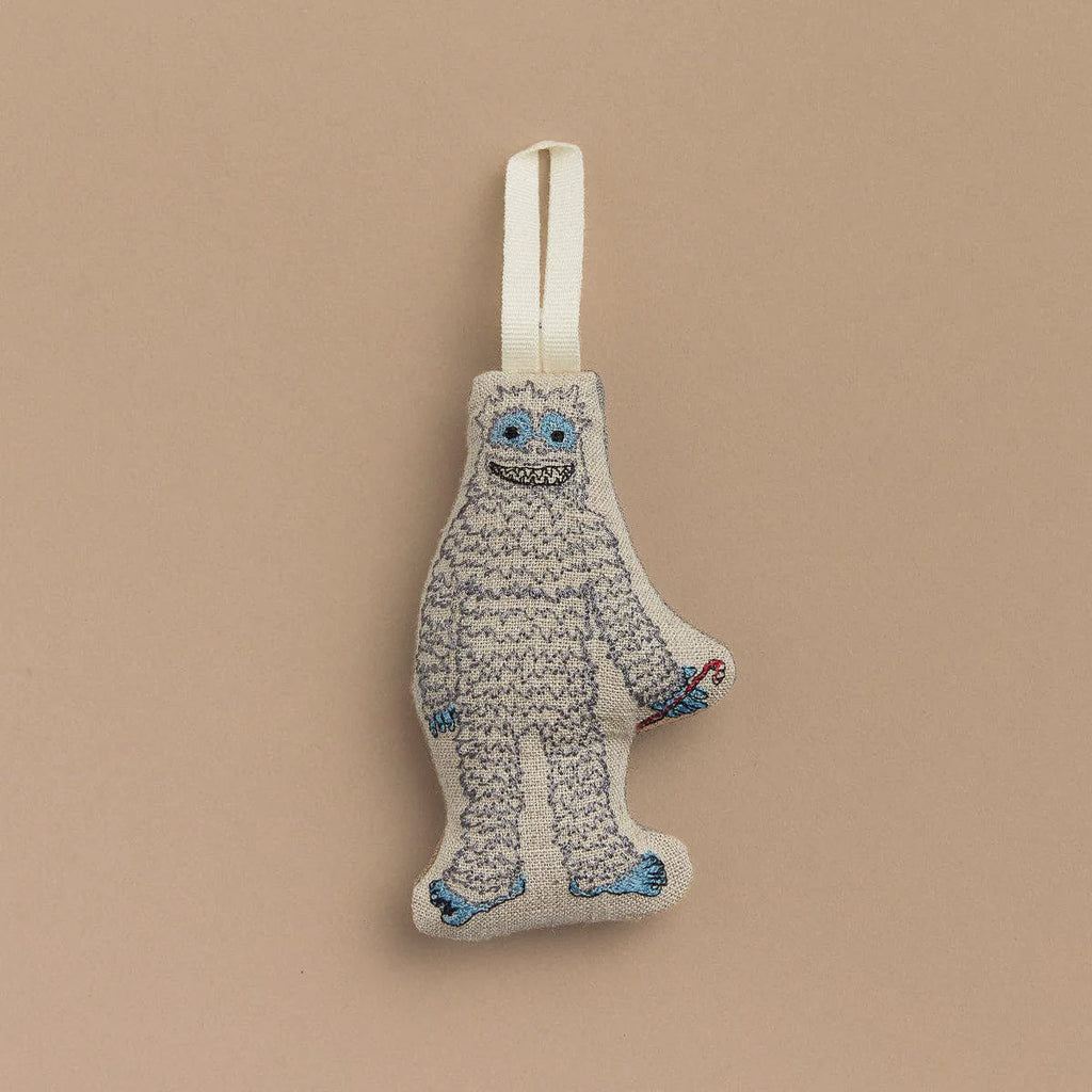 A Yeti with Candy Cane Ornament featuring a plush, fluffy monster with blue eyes and feet, hanging by a white ribbon against a beige background. This whimsical decoration makes the perfect gift for any occasion.