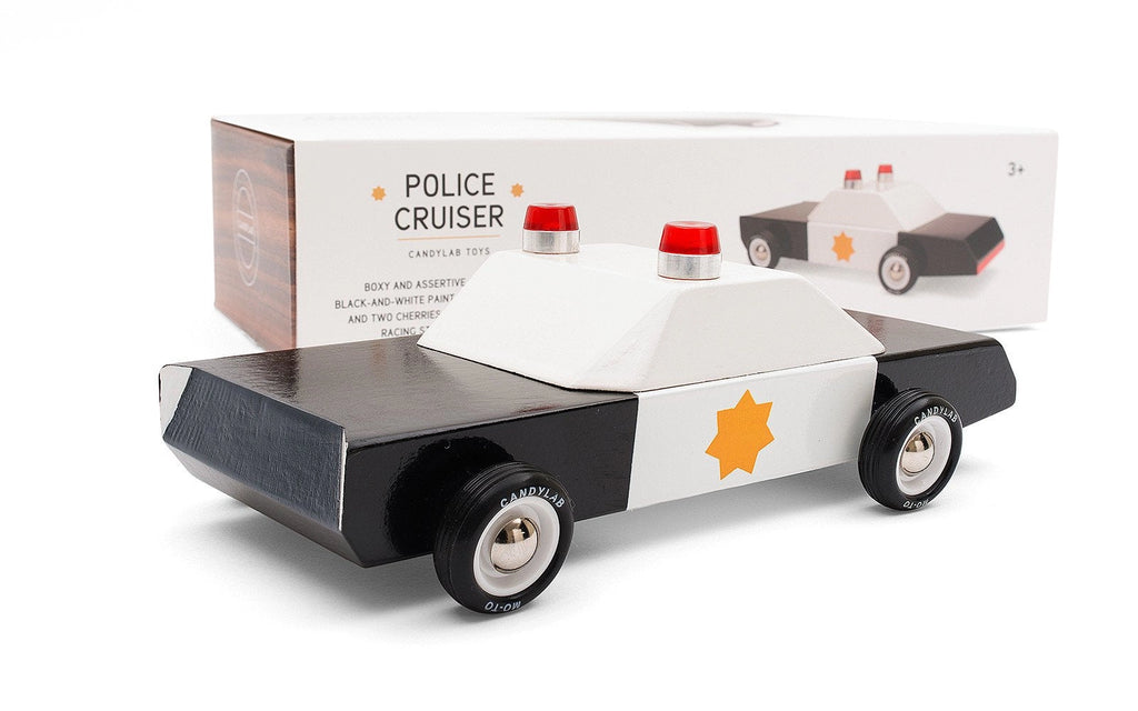 The Candylab Police Cruiser is a black and white wooden toy police car crafted from solid beech wood, showcasing a star emblem on its side, rubber tires, and two red lights on top. The toy is shown in front of its packaging, which features an image of the same safety-tested police cruiser.