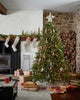 A decorated Christmas tree stands beside a stone fireplace, adorned with North Pole Bear Ornaments and lit with warm lights. Wrapped gifts are placed under the tree, and stockings hang on the mantel. A Santa Bear sits cheerfully among the presents, creating a cozy holiday scene.