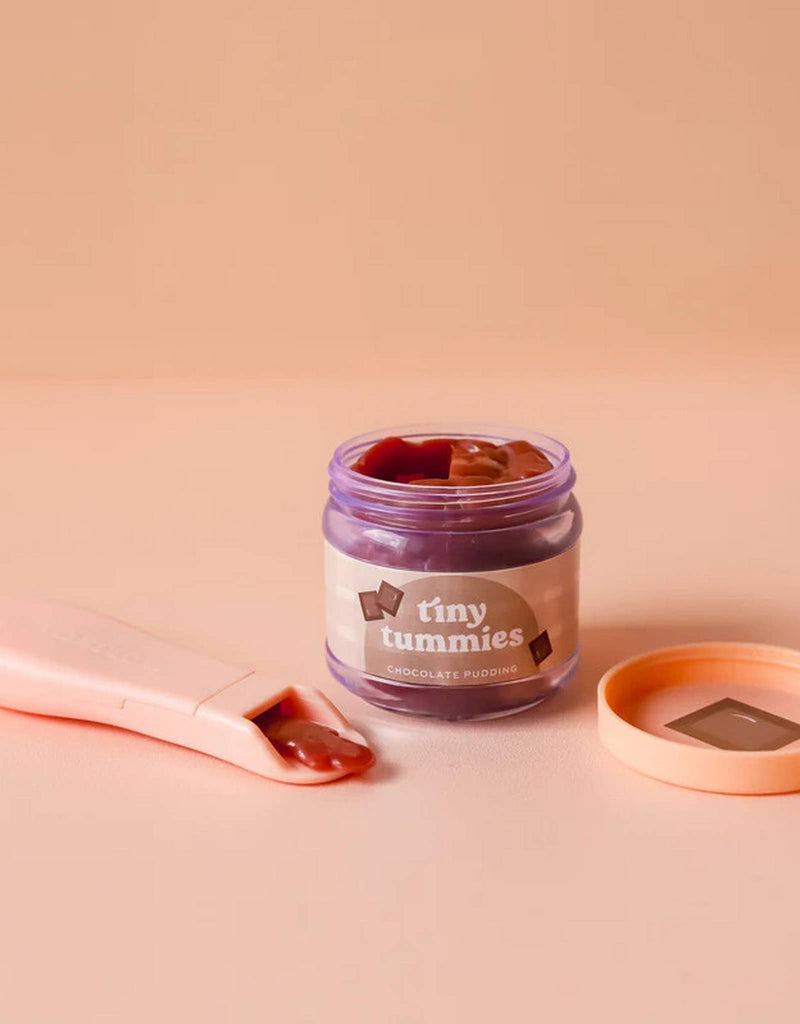 A jar of Tiny Tummies pudding from the Pink Gift Box - The Essentials by Tiny Harlow sits open, revealing rich chocolate inside. The orange lid rests nearby, with a spoonful of the delightful treat awaiting on a peach-colored background—perfect for doll essentials or a sweet addition to your gift box.