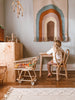 A young child sits at a small wooden table in a cozy room, facing a colorful woven rainbow tapestry on the wall. A miniature shopping cart filled with Toddler Rattan Bunny Chairs is beside them.