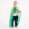 A joyful child stands barefoot, wearing the Forest version of Sarah's Silk Enchanted Playsilk as a green cape over a white shirt and dark pants. Engaged in imaginative play, they smile widely against the plain, light-colored background.