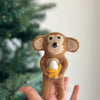 A hand holds a small "Monkey Holding Banana Finger Puppet" needle felted wool figurine in front of a blurred Christmas tree. The monkey, crafted from 100% wool, is brown with light-colored inner ears and belly, clutching a yellow and white banana. This handmade finger puppet stands out against the softly out-of-focus background.